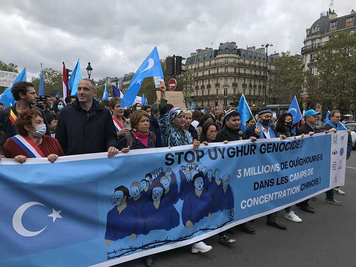 Glucksmann en compagnie de l’écolo belge Cogolati derrière une pancarte mensongère aux couleurs du « Turkestan oriental ». Au milieu, la tête de Dilnur Reyhan.